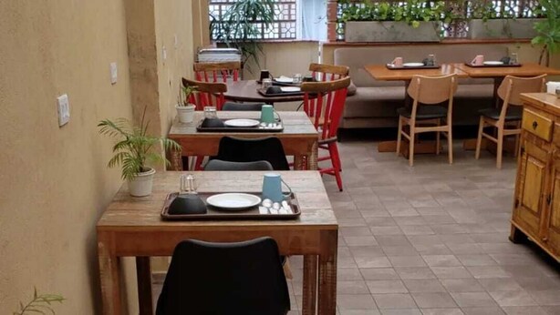 A dining area with wooden tables and chairs, potted plants, and a kitchen in the background. - Soul Hostel 2 (Sao Paulo)