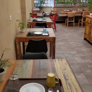 A dining area with wooden tables and chairs, potted plants, and a kitchen in the background. - Soul Hostel 2 (Sao Paulo)