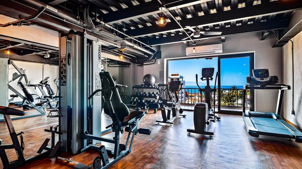 A gym with various exercise equipment, a large window with a view of the sea, and a ceiling with exposed pipes.