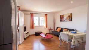 A compact living space with a kitchenette, a dining area with a table and chairs, a sofa, and a window with curtains. - Llevant 11 E1 (Cap d'Artrutx)