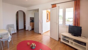 A living room with a TV, a red table, and a kitchen in the background. - Llevant 11 E1 (Cap d'Artrutx)