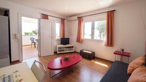 A spacious living area with a sofa, a red table, a television on a white cabinet, and a suitcase. - Llevant 11 E1 (Cap d'Artrutx)