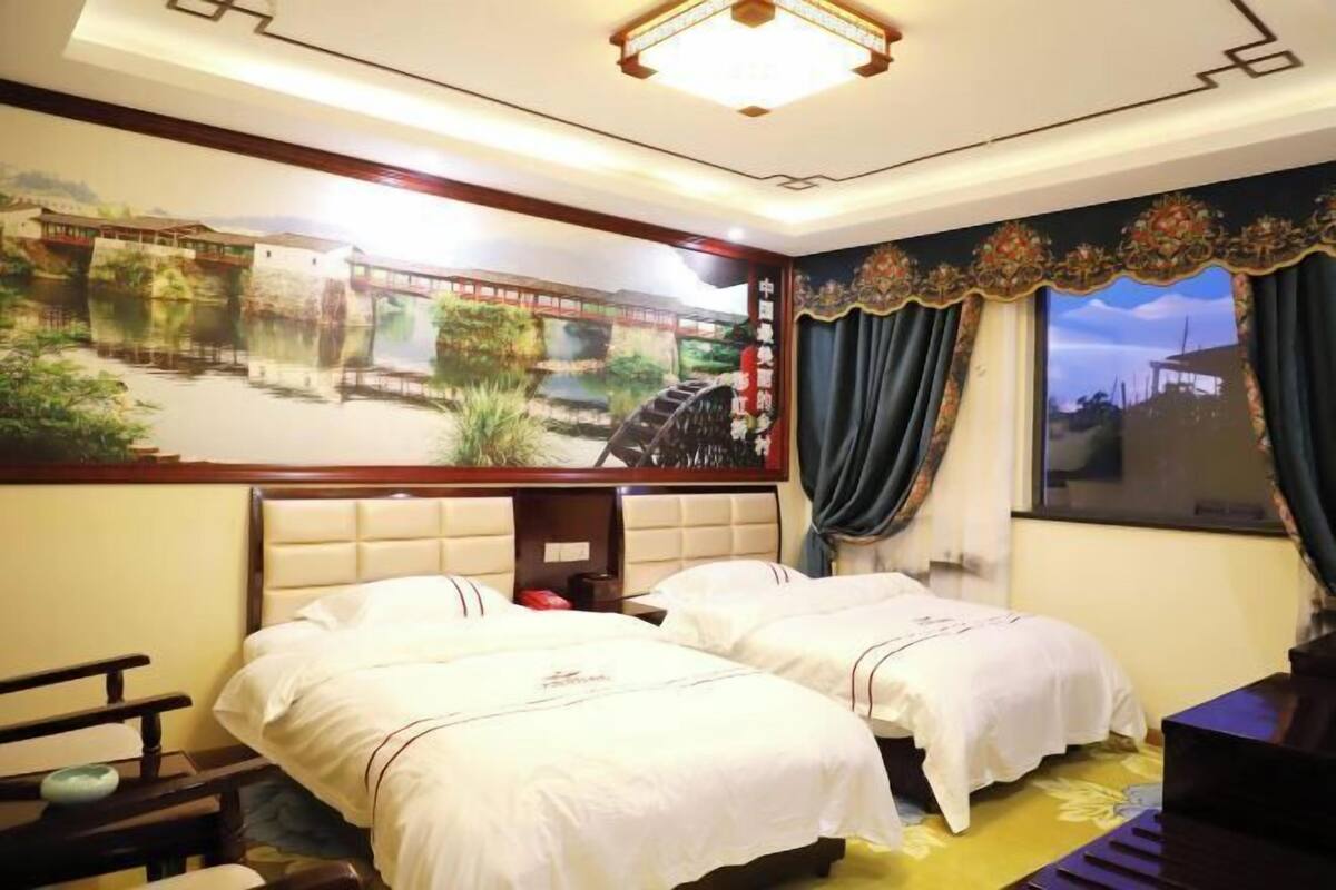 A hotel room with two beds, a large mural of a bridge over water, a desk with a chair, and a view of the outside through a window with curtains.