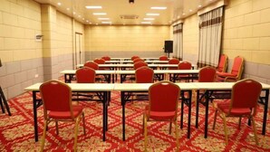 A conference room with long tables and red chairs, carpeted floor, and a plain wall with a door and a speaker.