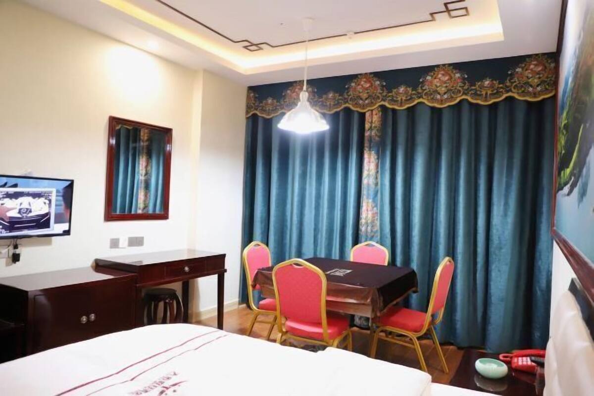 A hotel room with a bed, a desk, a television, a dining table with chairs, and a large window with curtains.