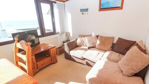Una sala de estar acogedora con un sofá, un mueble de televisión de madera, una mesa y vistas al mar a través de un ventanal amplio.