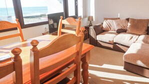 Un salón acogedor con una mesa de comedor de madera, sillas, un sofá y vistas al mar a través de amplios ventanales.