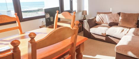 A cozy living room with a wooden dining table, chairs, a sofa, and a view of the sea through large windows.