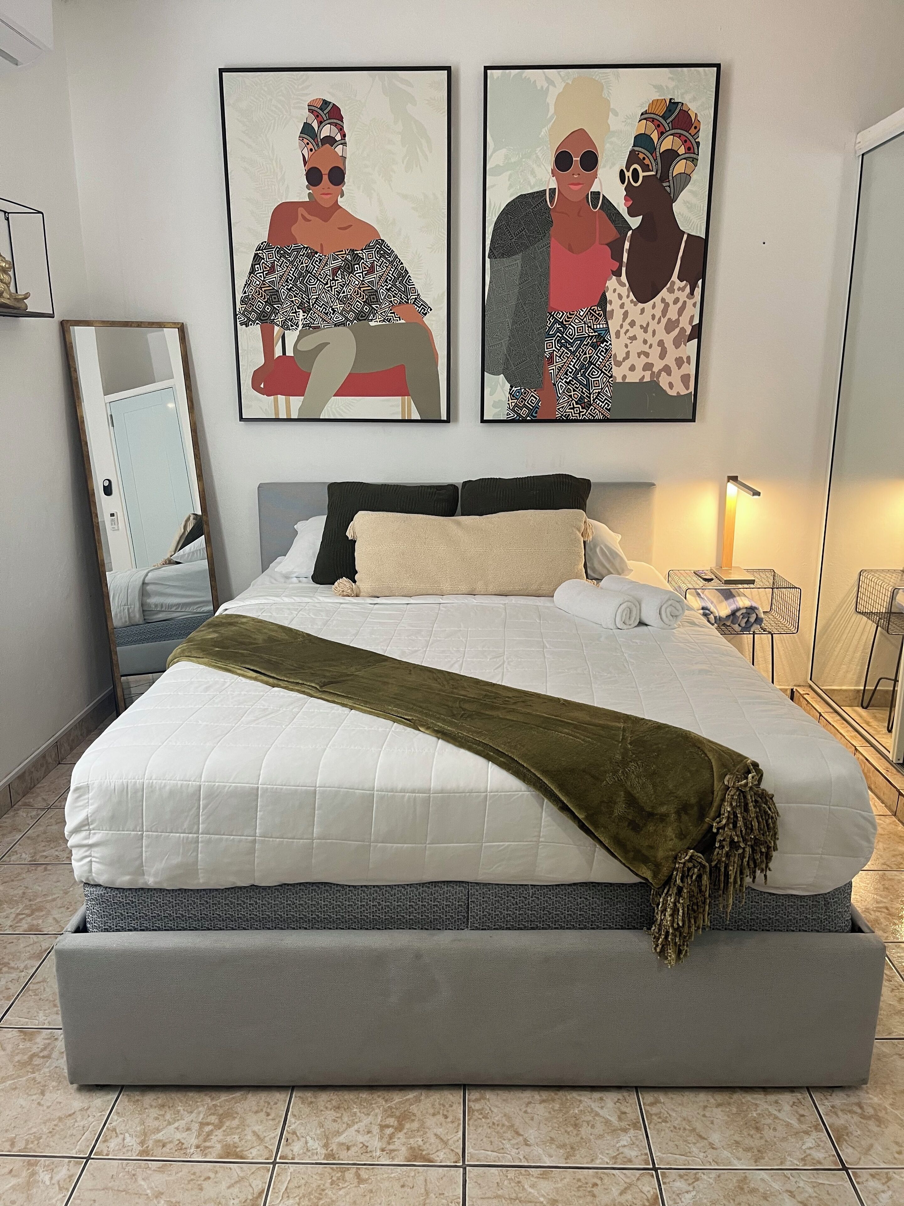Uma cama bem arrumada com colcha branca e manta verde, uma mesinha de cabeceira com abajur e duas obras de arte emolduradas na parede.