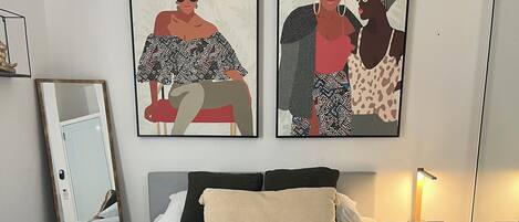 Uma cama bem arrumada com colcha branca e manta verde, uma mesinha de cabeceira com abajur e duas obras de arte emolduradas na parede.