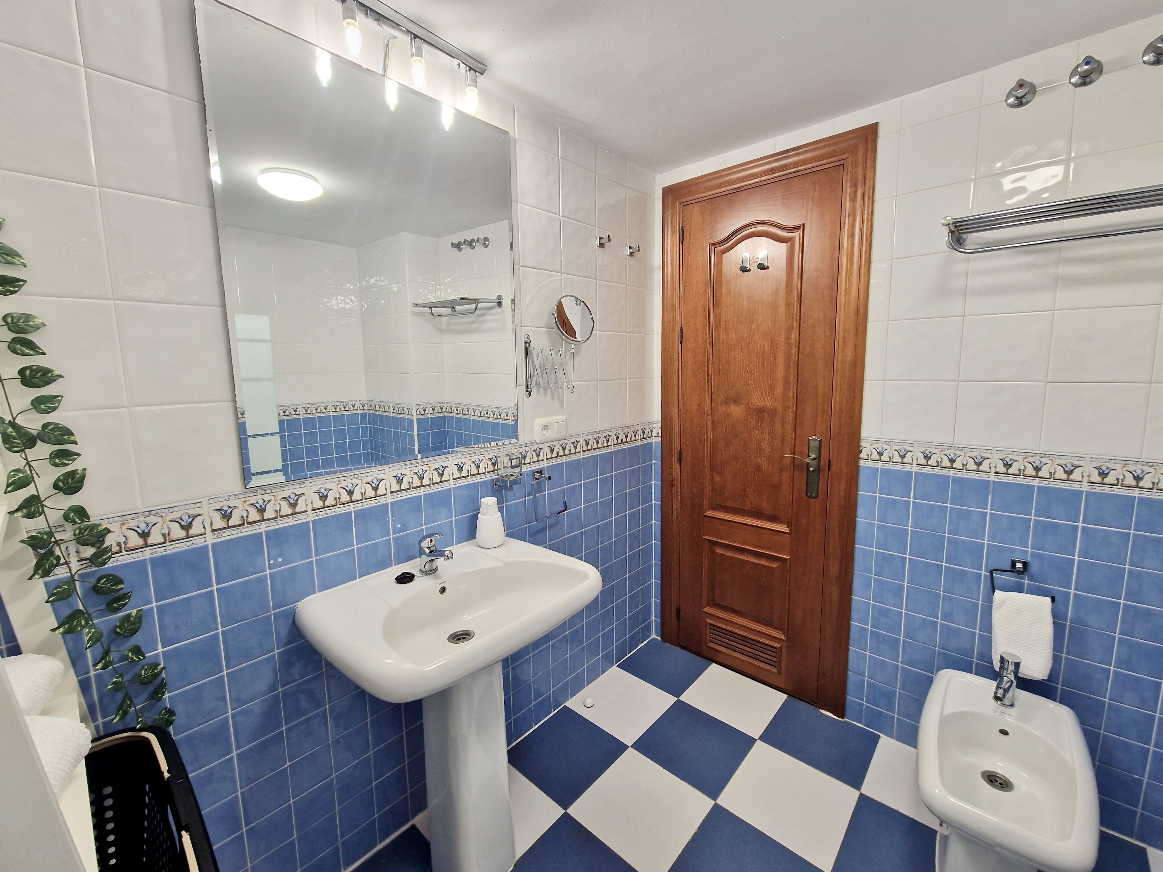 Un baño con suelo de baldosas azules y blancas, una puerta de madera, un lavabo blanco, un bidé y un espejo con iluminación.