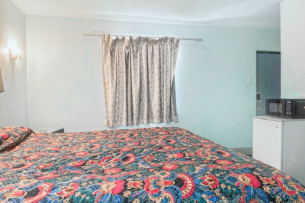 A bedroom with a patterned bedspread, a hanging curtain, a white wall, and a small white cabinet with a microwave.