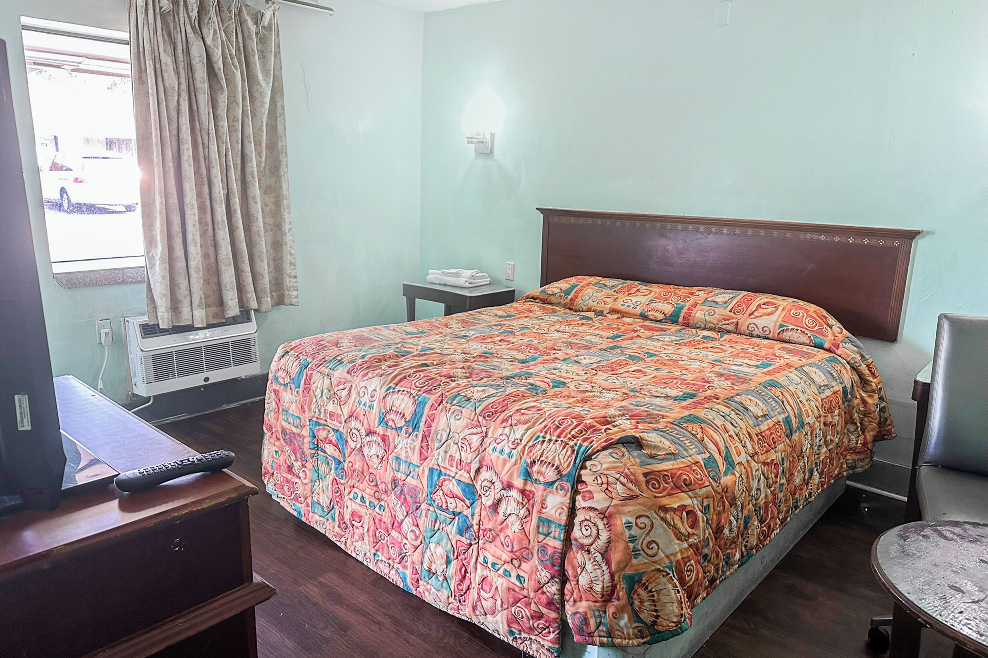 A bedroom with a bed, a desk, a chair, a window with curtains, and an air conditioning unit.