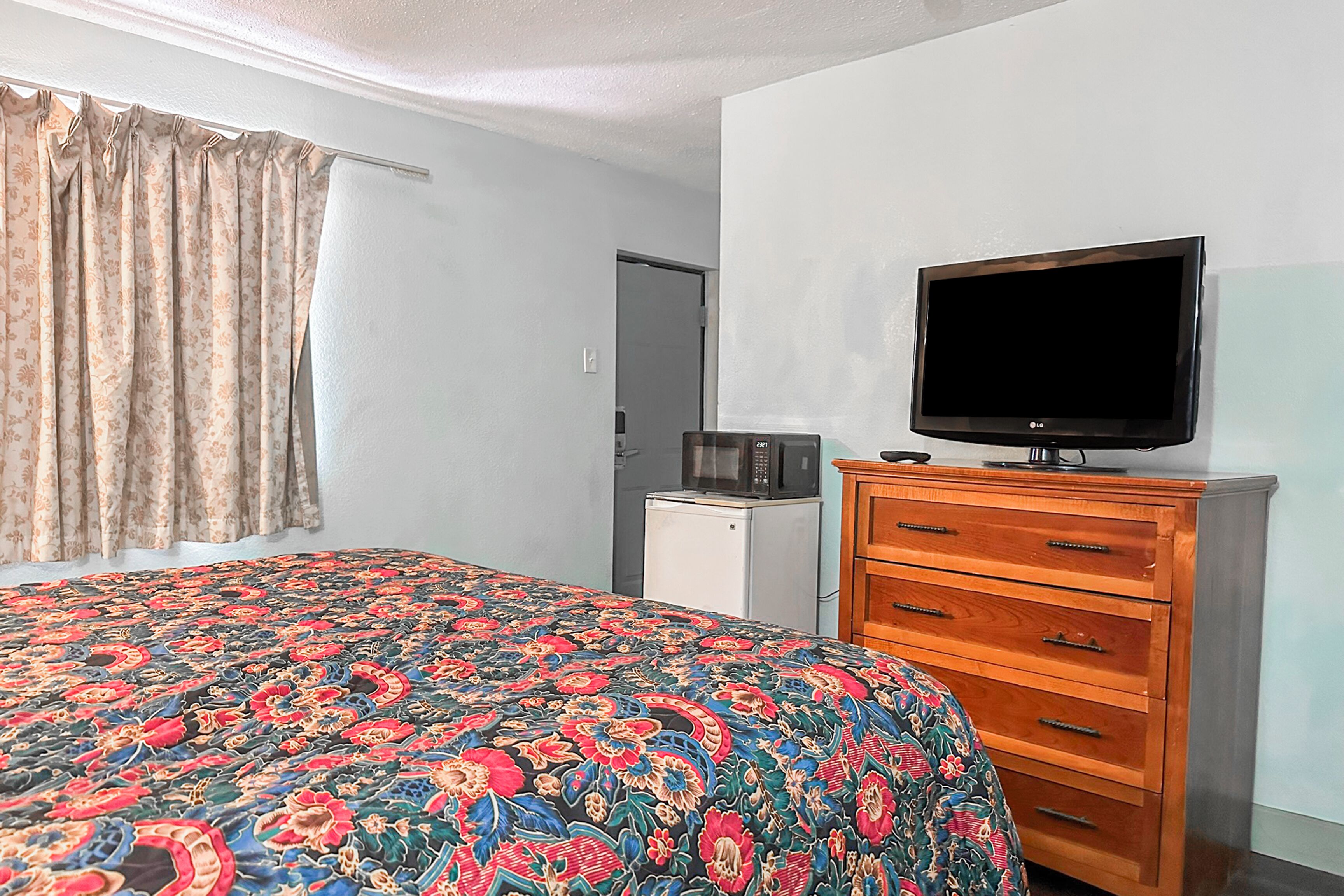 Quarto com uma cama, televisão sobre uma cômoda de madeira, micro-ondas e uma porta.