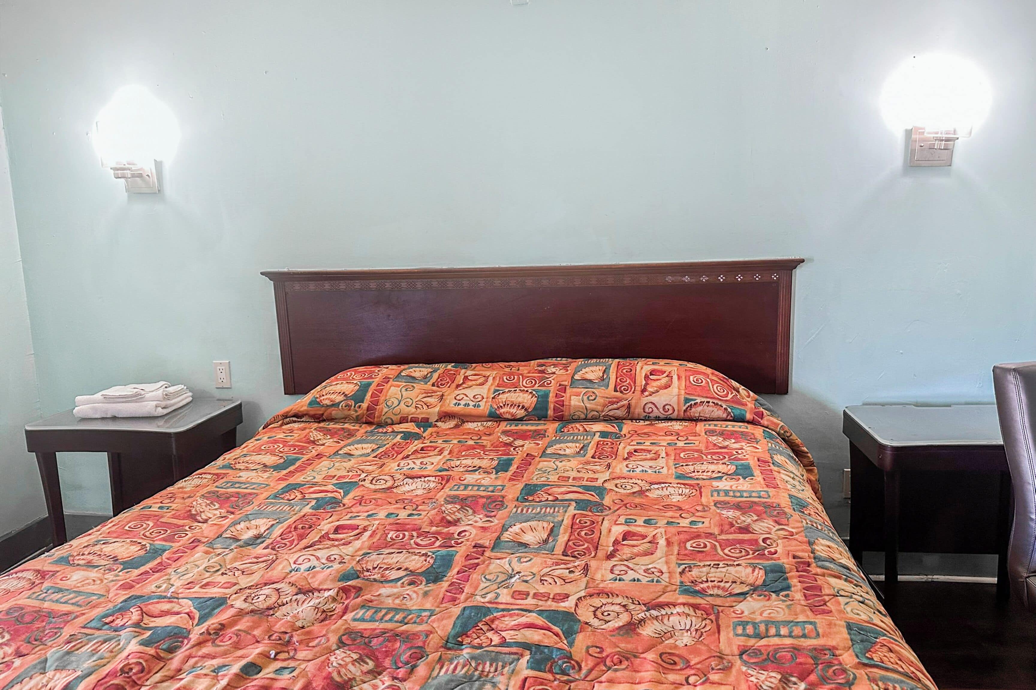 Quarto com cabeceira de madeira, colcha estampada, duas mesas de cabeceira e luminárias fixadas na parede.