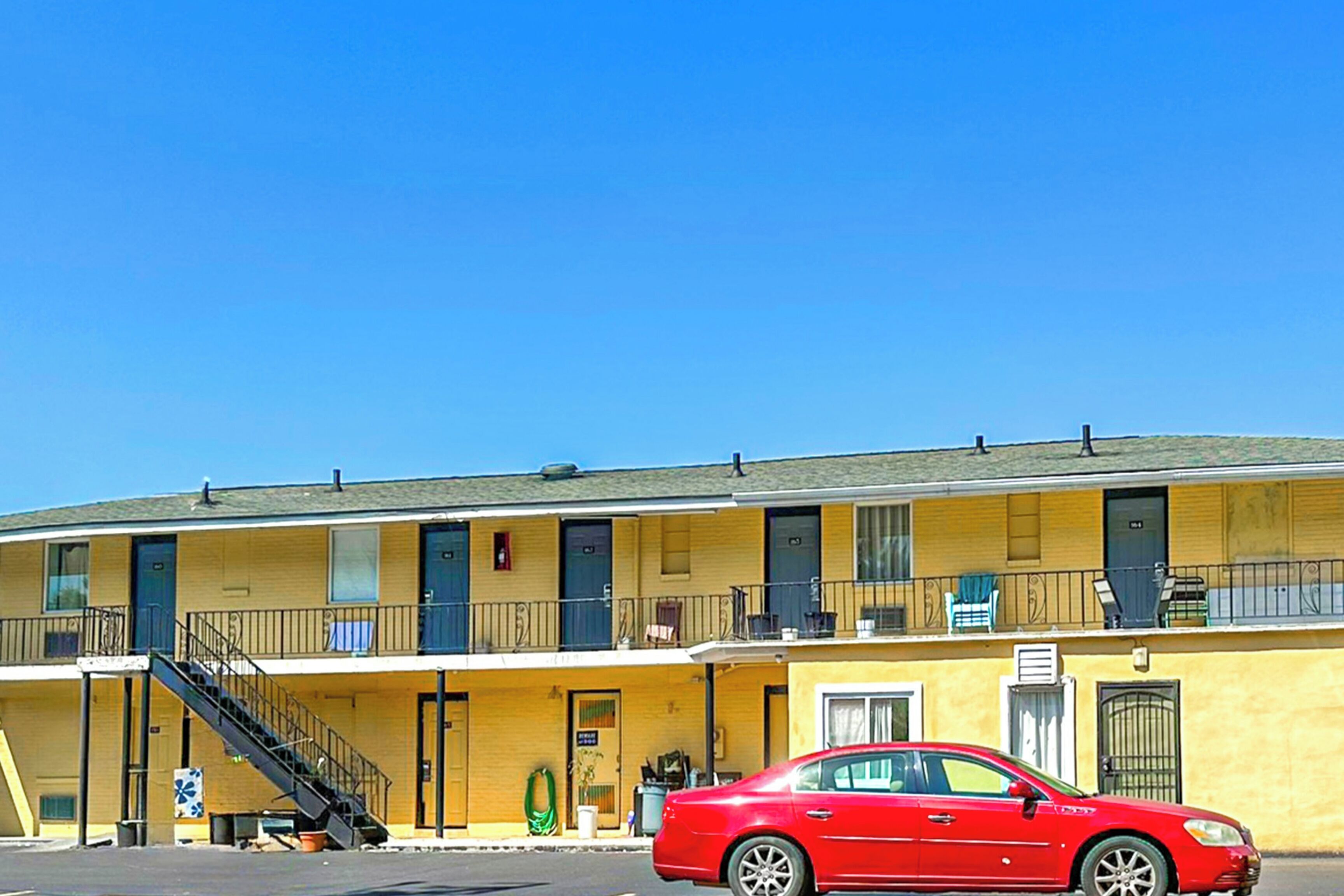 A motel with a red car parked in front. It has multiple rooms with balconies and a staircase leading to the entrance.