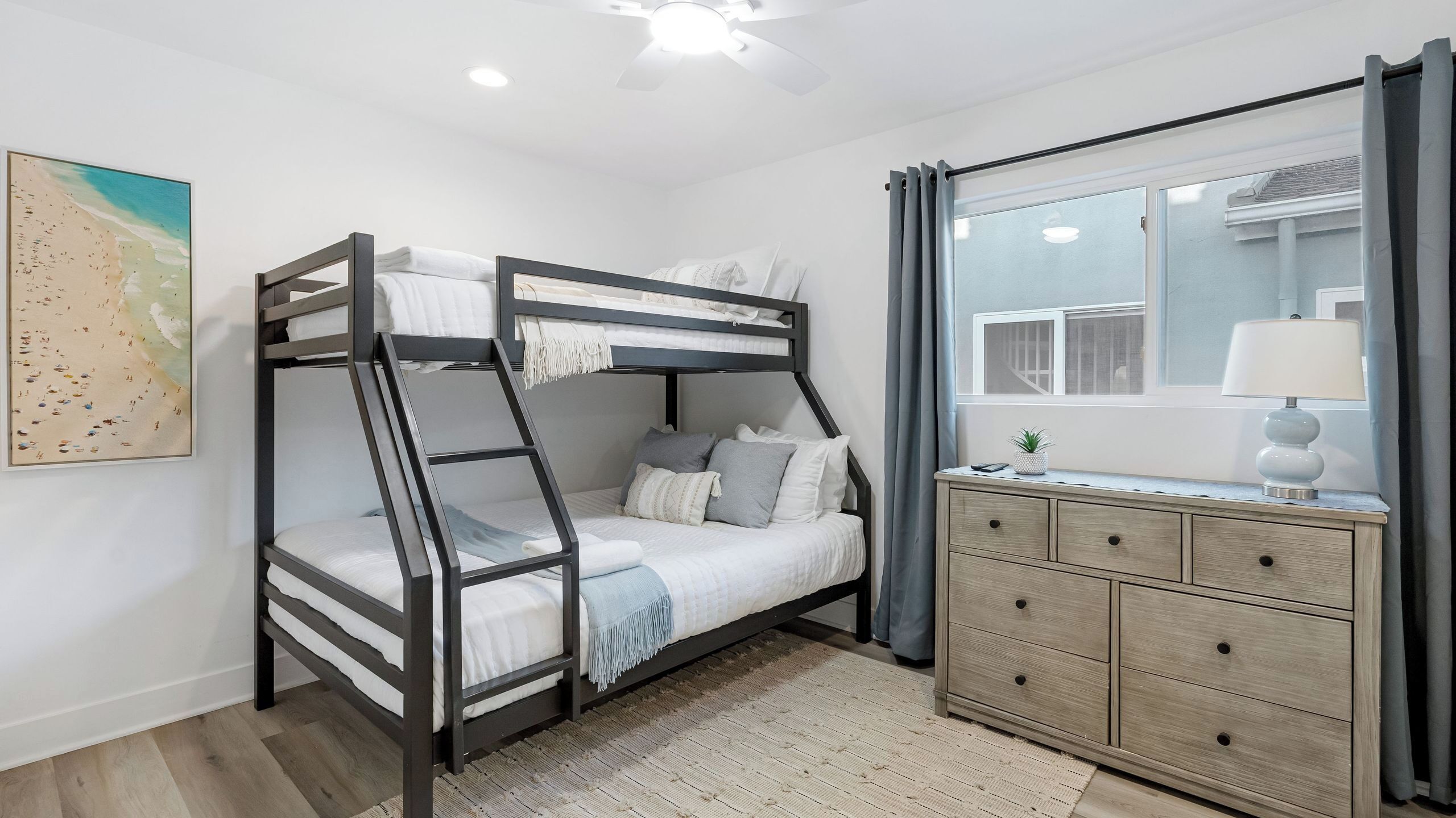 A bunk bed with a desk and drawers, a window with curtains, and a beach-themed painting on the wall.