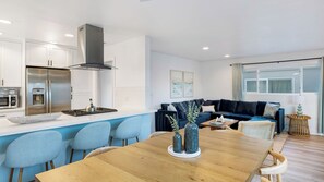 A modern kitchen with a central island, a dining area with a wooden table, and a living room with a dark blue sofa.
