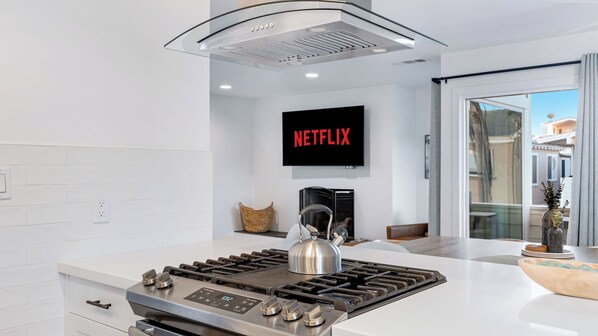 A modern kitchen with a gas stove, a kettle, and a wall-mounted television displaying 'NETFLIX'.