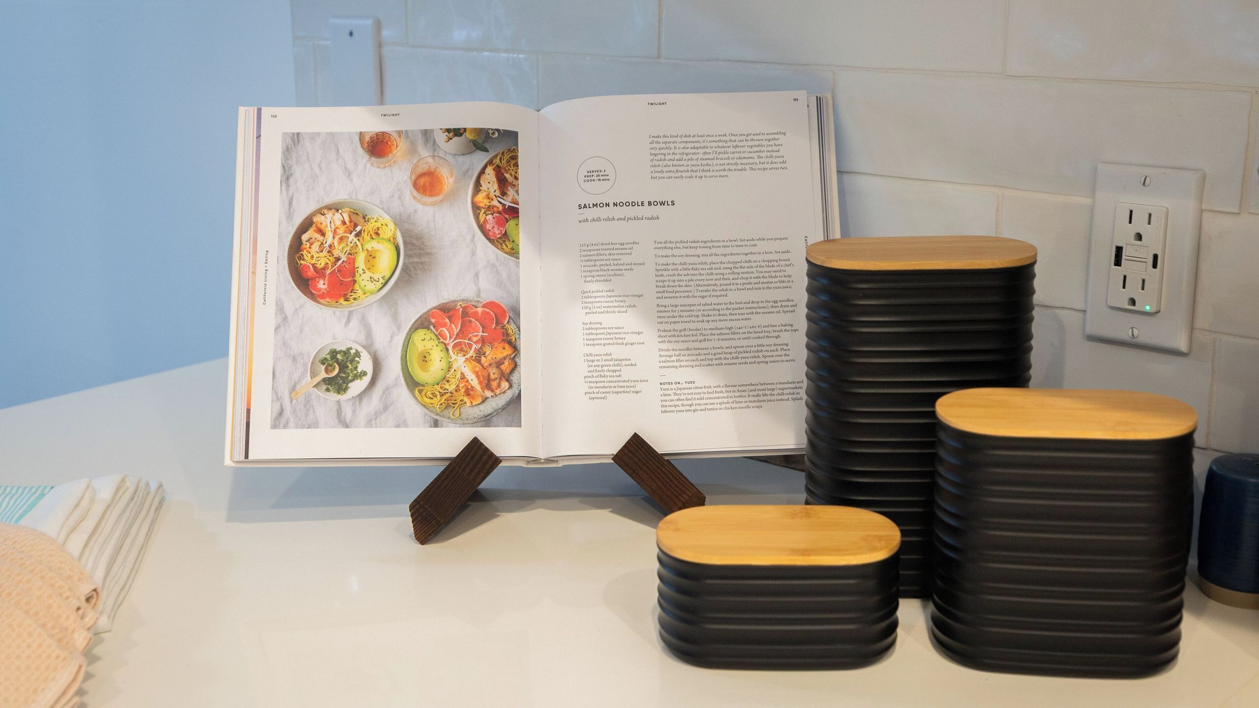 Un libro de cocina abierto sobre una encimera con pilas de tazones negros y una cuchara de madera.