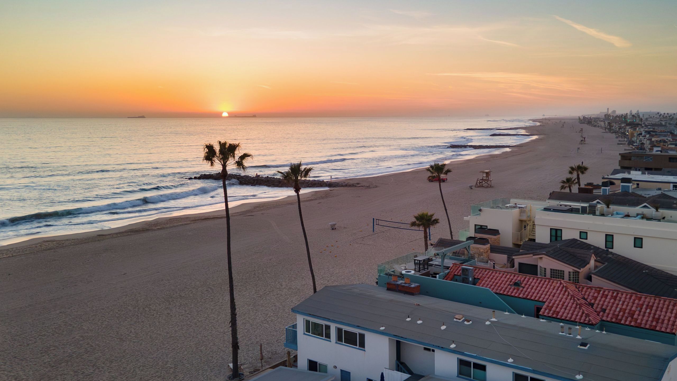 A beach view with a sunset, palm trees, and buildings with red roofs.
