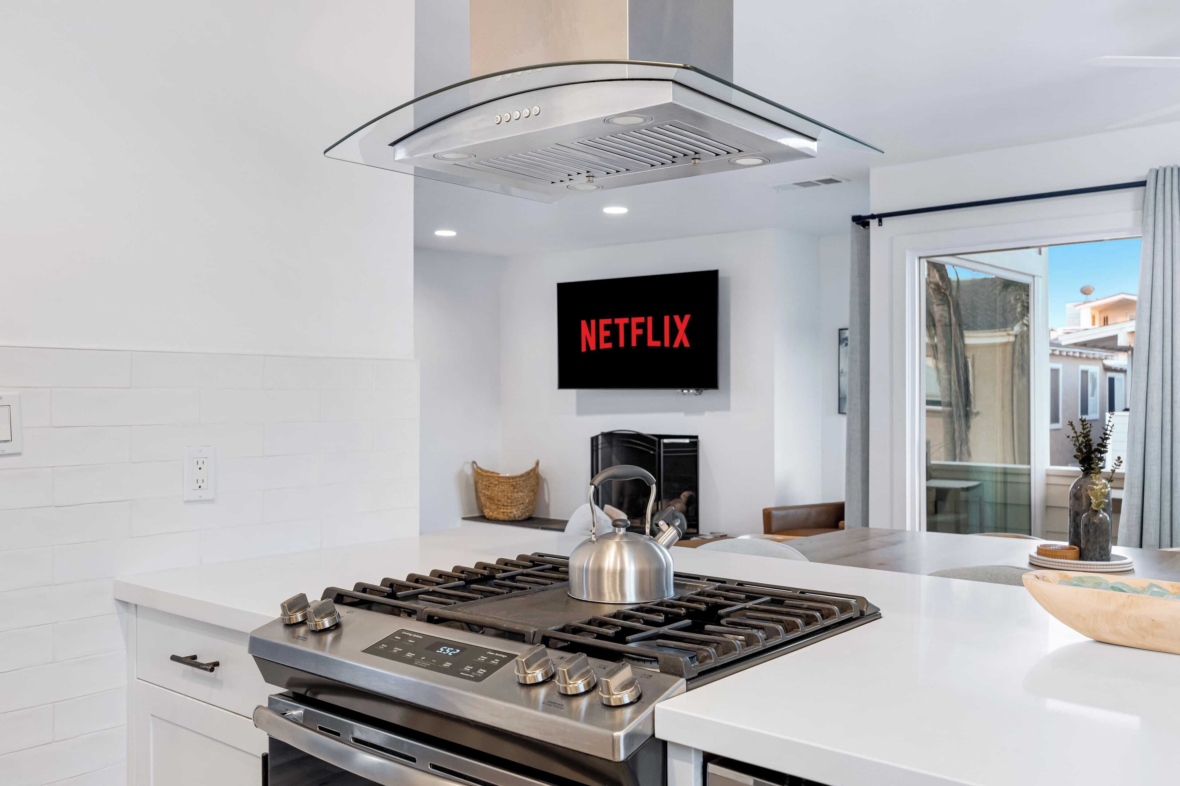 A modern kitchen with a gas stove, a kettle, and a flat-screen TV displaying "NETFLIX".