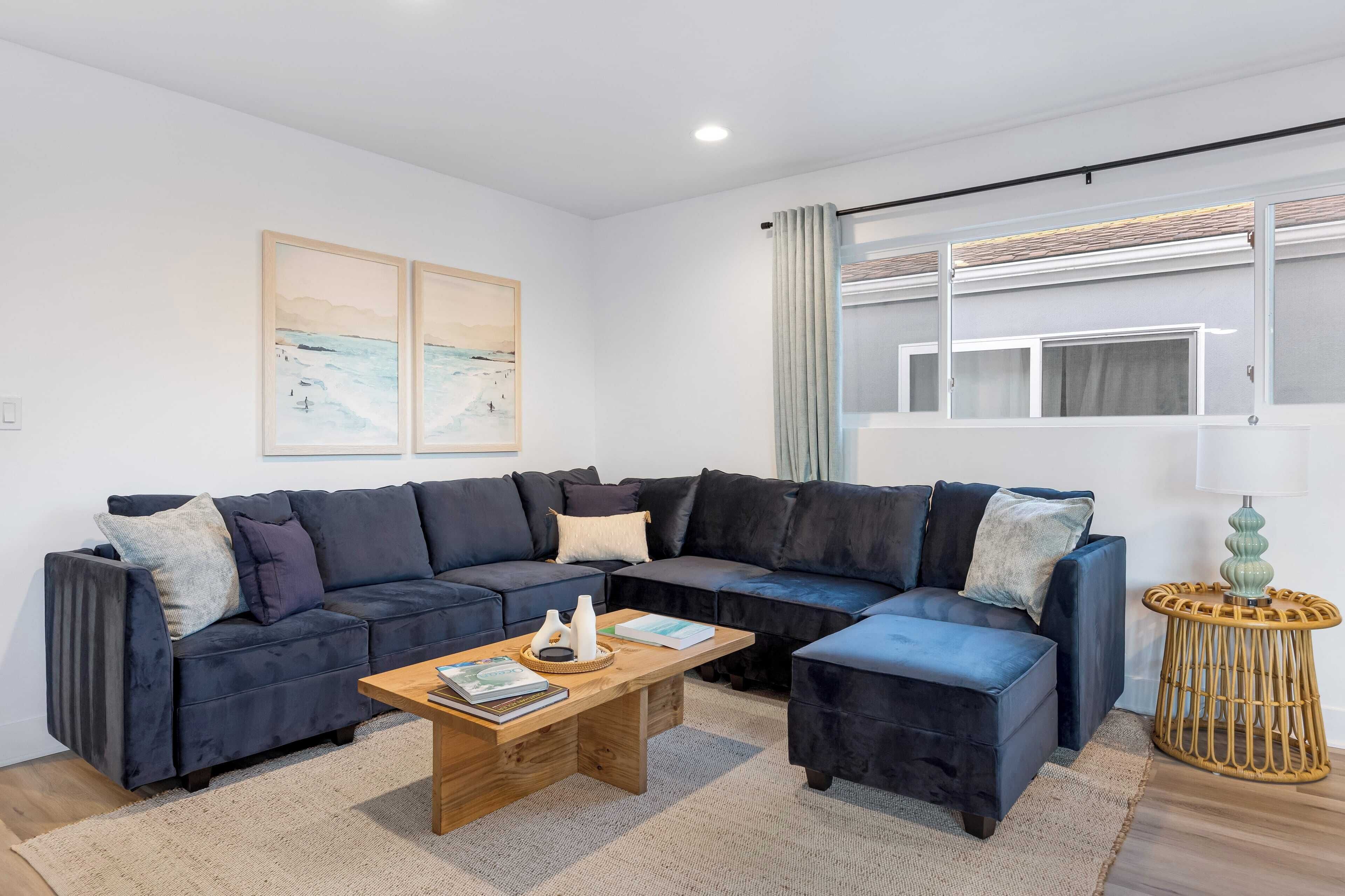 A modern living room with a dark blue sectional sofa, a wooden coffee table, and a small side table with a lamp. There are two framed artworks on the wall and a window with curtains.