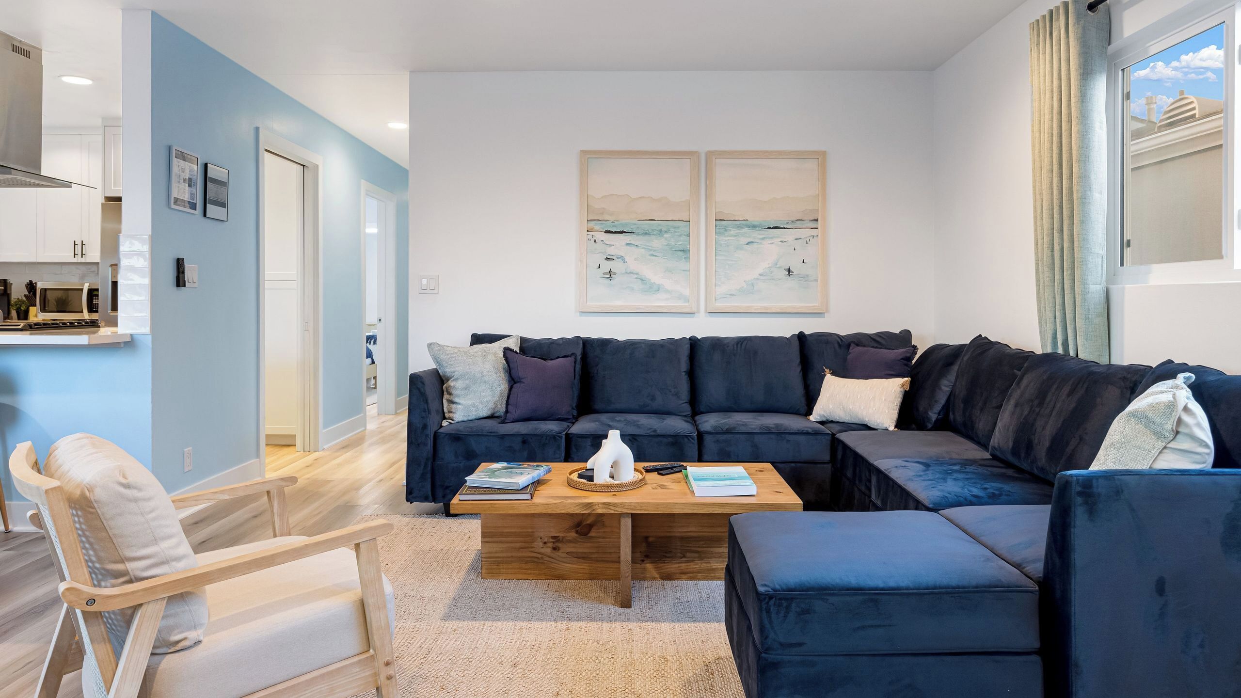Una sala de estar moderna con un sofá seccional azul oscuro, una mesa de centro de madera y un sillón de color claro. Hay dos cuadros enmarcados en la pared y una ventana con cortinas.