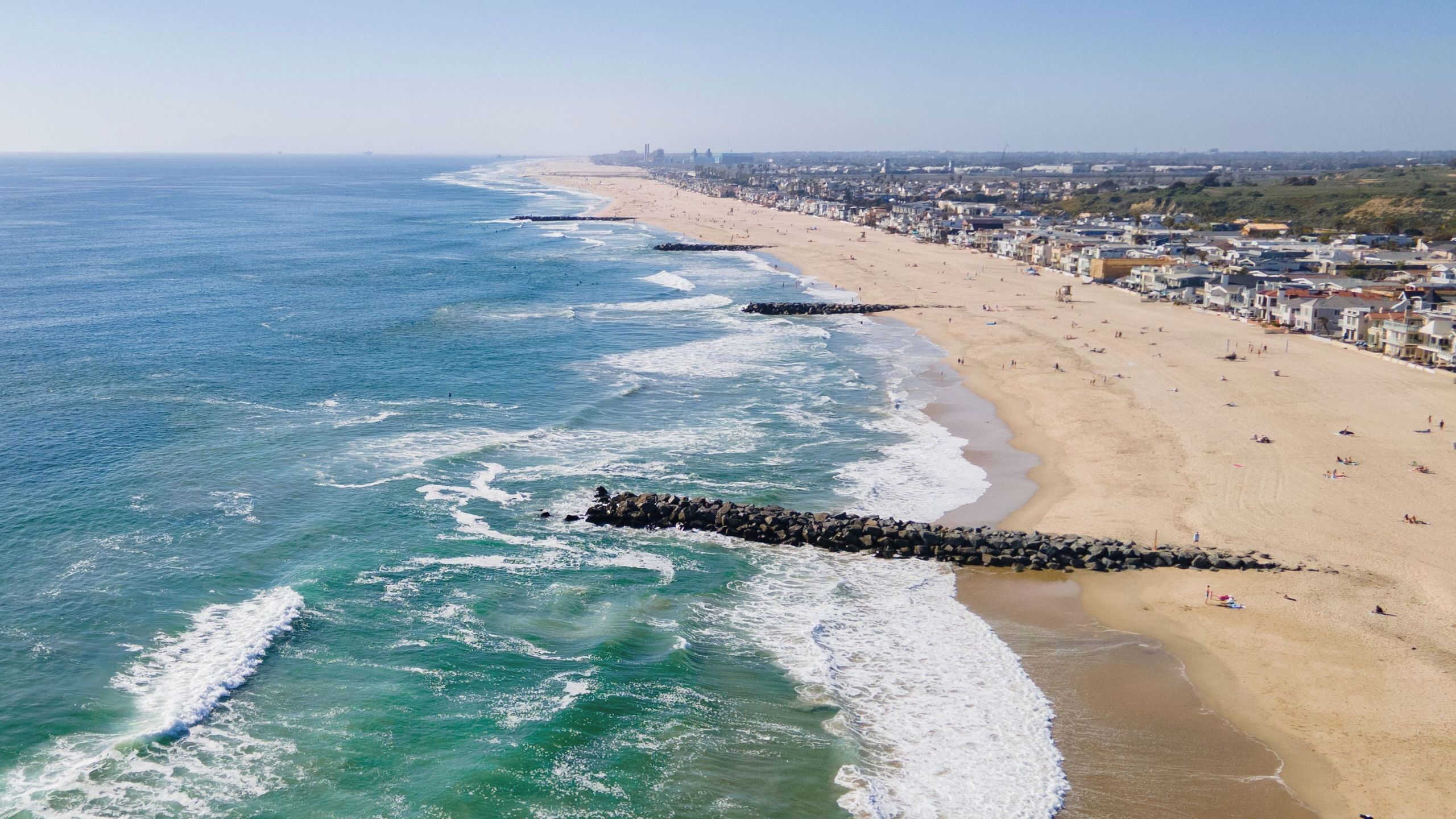 Una playa costera con un rompeolas rocoso, una orilla arenosa y un paisaje urbano al fondo.
