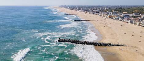 Una playa costera con un rompeolas rocoso, una orilla arenosa y un paisaje urbano al fondo.