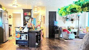 A cozy living room with a wooden floor, a central island with various items, a large tree, and a door leading to another room. - Whimsical 3 bd cottage for families and pets. Sleeps 7, free wifi/snacks (Midwest City)