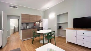 Une cuisine moderne avec des meubles en bois, un réfrigérateur blanc et une cuisinière noire. On y trouve un coin repas avec une table blanche et des chaises vertes. Un téléviseur à écran plat est fixé au mur.