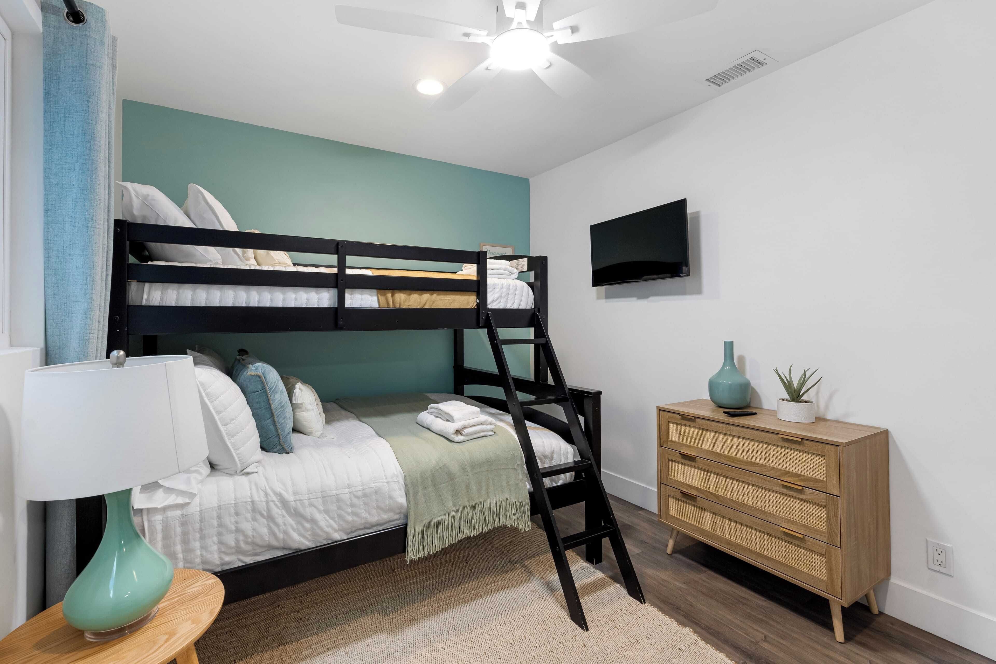Un lit superposé avec une échelle, une table de chevet avec une lampe, une commode et un téléviseur fixé au mur.