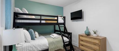 Un lit superposé avec une échelle, une table de chevet avec une lampe, une commode et un téléviseur fixé au mur.