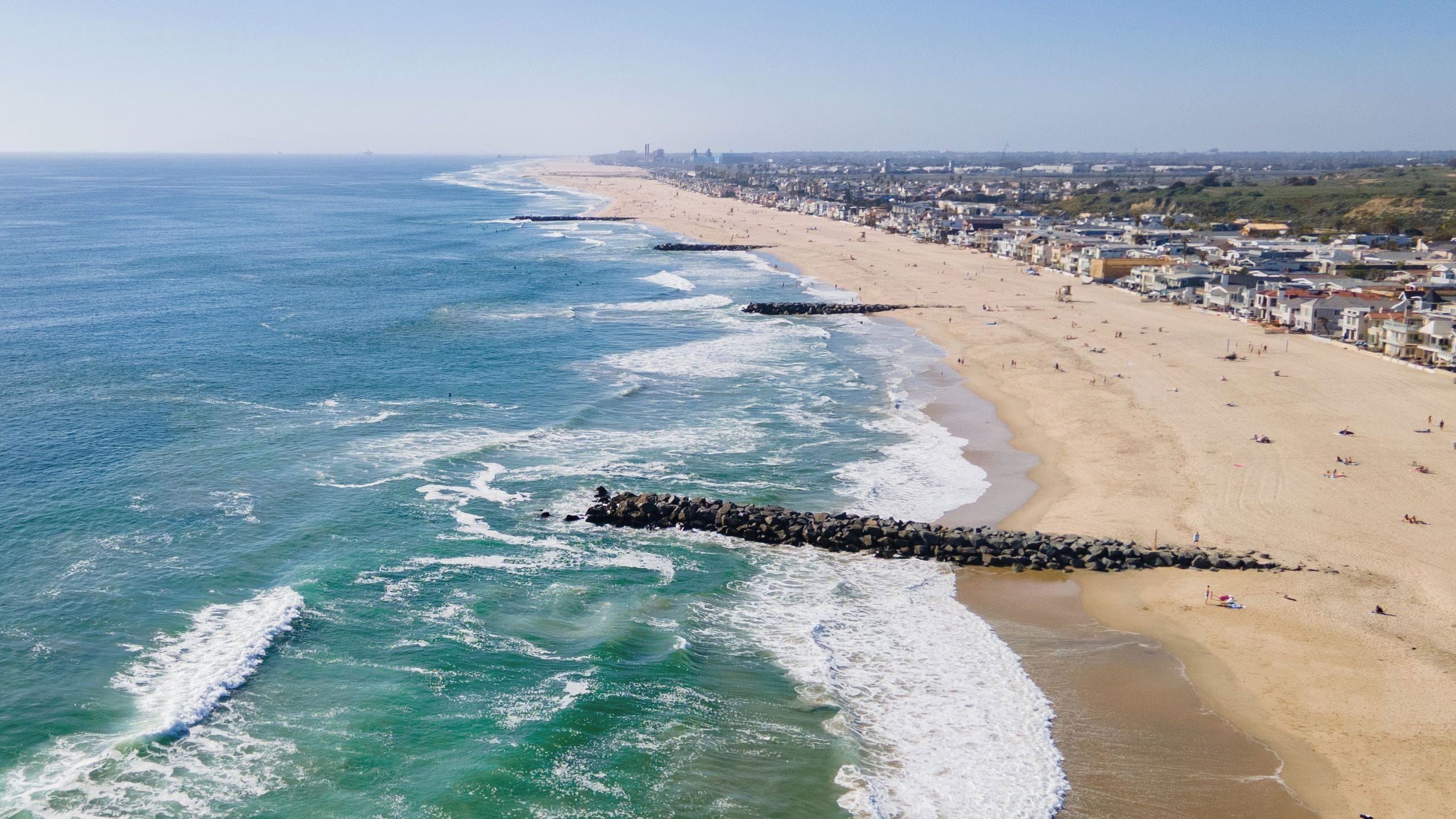 Una playa costera con un rompeolas rocoso, una orilla arenosa y un paisaje urbano al fondo.