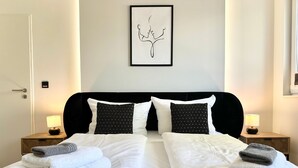 Un lit bien fait, avec du linge de lit blanc et deux coussins noirs à motifs, flanqué de deux tables de chevet avec des lampes, dans une pièce au design simple et épuré.