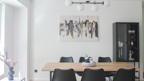 Un espace repas moderne avec une table en bois, des chaises noires, un meuble vitré et un tableau représentant un paysage urbain accroché au mur.