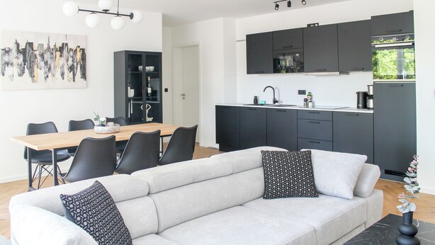 Un salon moderne avec un canapé et des fauteuils, un coin repas avec une table et des chaises, et une cuisine avec des meubles et des appareils électroménagers aux couleurs sombres.