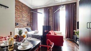 A modern hotel room with a brick wall, a red sofa, a dining table with a bottle of wine, and a bed with a view of the city. - Cracow Fancy Brick #1 – Old Town (Kraków)