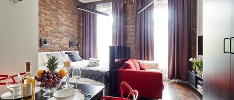 A modern hotel room with a brick wall, a red sofa, a dining table with a bottle of wine, and a bed with a view of the city.