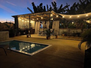 Un área de piscina con zona de estar cubierta, plantas en macetas y un bar.