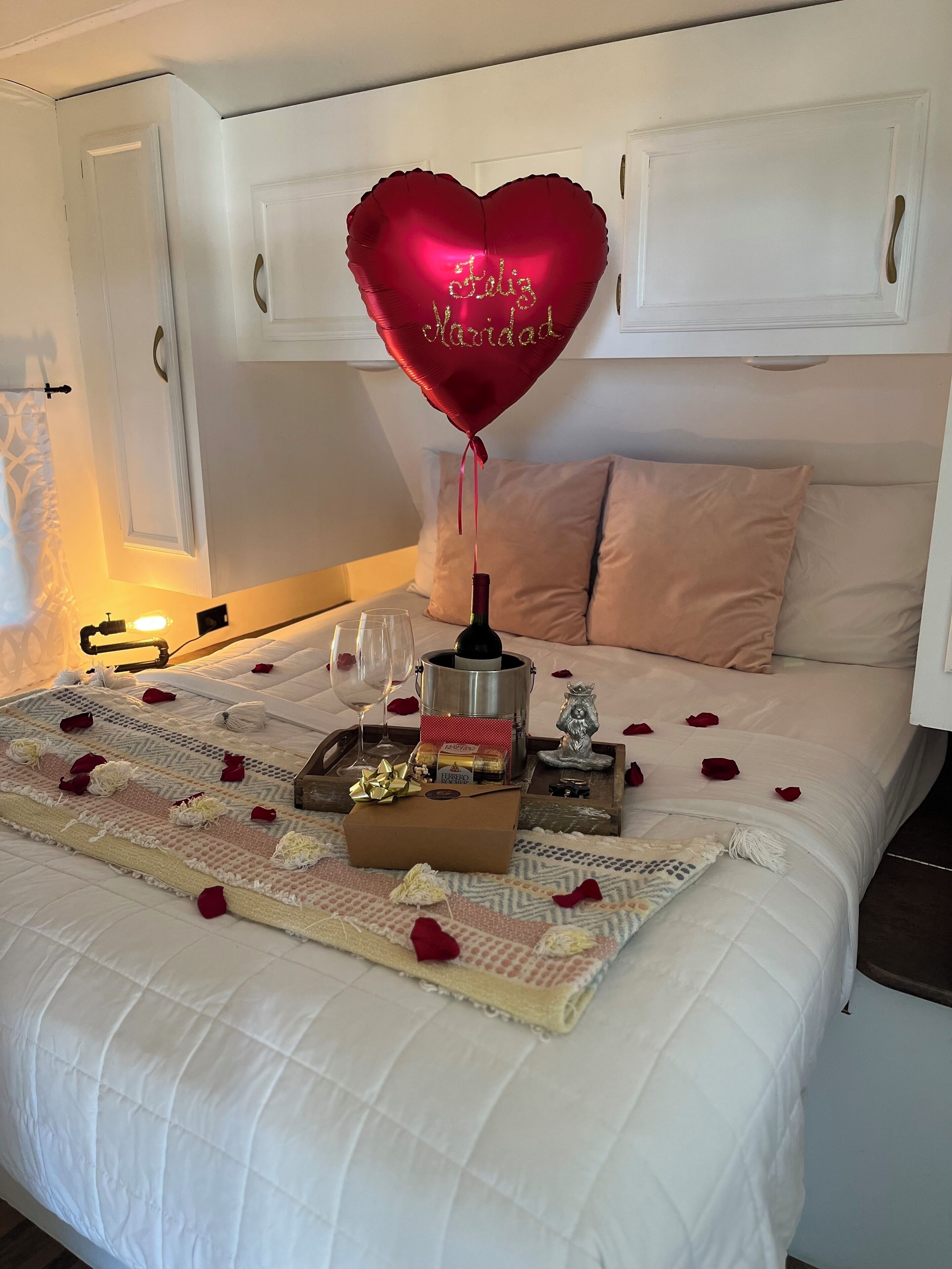 Una cama bien hecha adornada con un globo en forma de corazón, pétalos de rosa y una botella de champán.