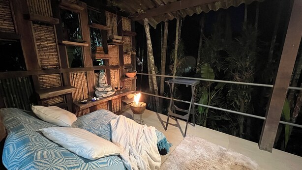 A room with a bed, a nightstand, a small table with a candle, and a view of the outdoors.