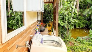 A compact kitchen with a sink, a small table, and a chair. There is a window with a view of greenery and a water body outside.