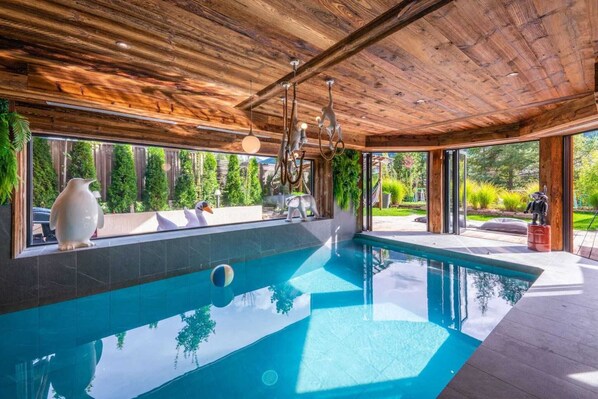 A modern indoor swimming pool with wooden ceiling beams, large windows, and outdoor views.
