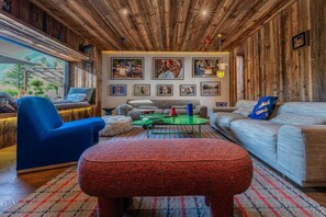 A cozy living room with wooden walls, a blue armchair, a grey sofa, and a red ottoman.
