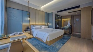 A modern hotel room with a large bed, bedside tables, a desk with a laptop, and a bathroom with a glass shower. - Wangfu International Hotel (Yiwu)