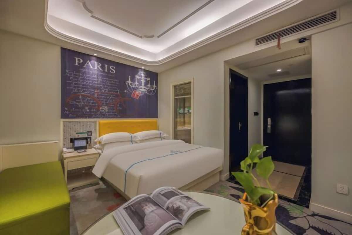 Habitación de hotel con cama, mesita de noche, un cuadro con temática parisina en la pared, una silla verde y una planta sobre una mesa.