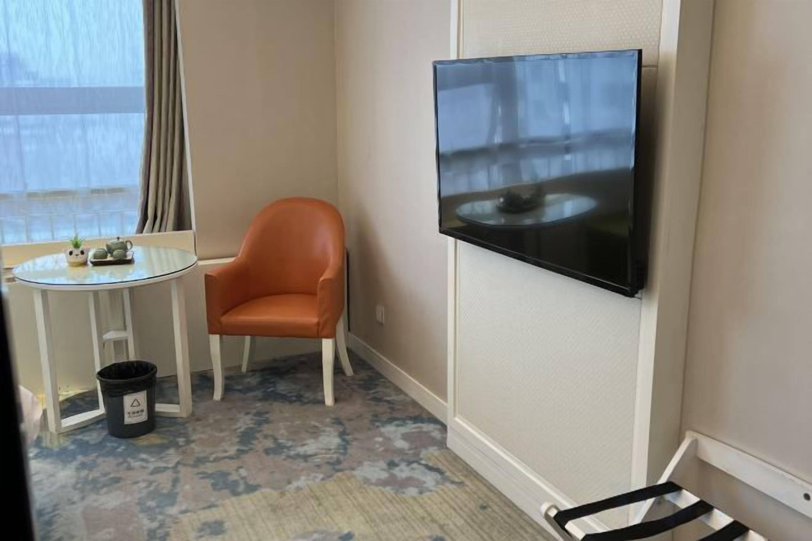 A hotel room with a television mounted on the wall, a small round table with a chair, and a window with curtains.