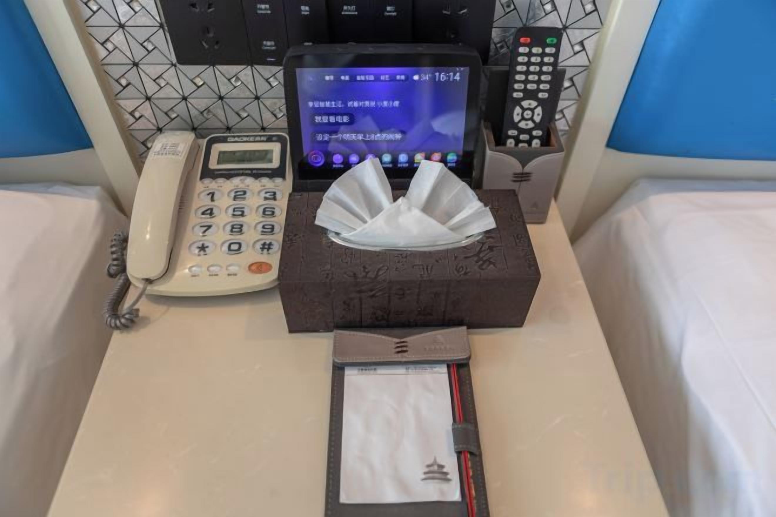 A hotel room with a bedside telephone, a digital clock, a tissue box, and a notepad on the bed.